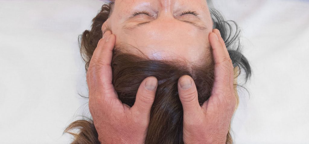 Cranio-Sacral-Behandlung Kopf einer liegenden Frau mit geschlossenen Augen von oben zu sehen, die Hände des Craniosacral-Spezialisten sind an ihren Schläfen.