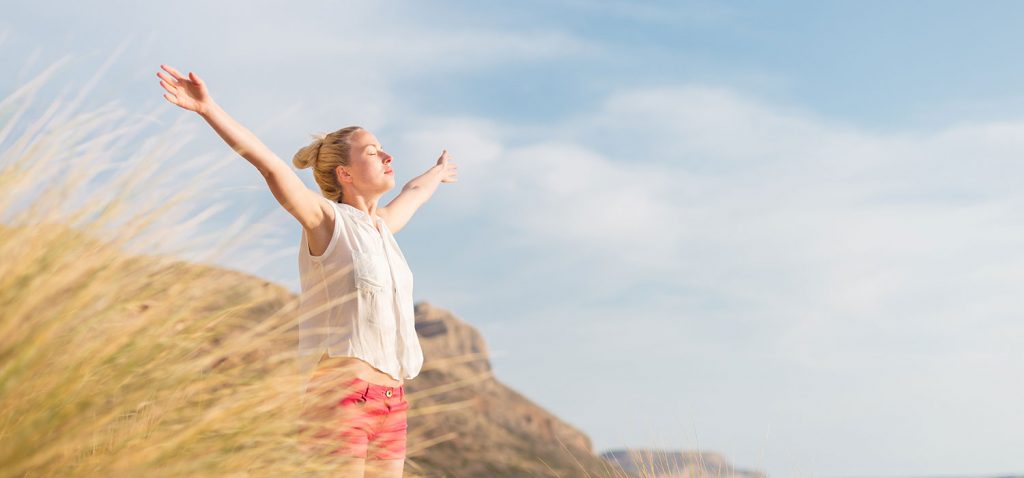 Frau in Dünen Frau, die mit ausgebreiteten Armen, geschlossenen Augen und sichtlichem Wohlbefinden vor blauem Himmel in einer Dünenlandschaft steht. - Matej Kastelic/Shutterstock.com