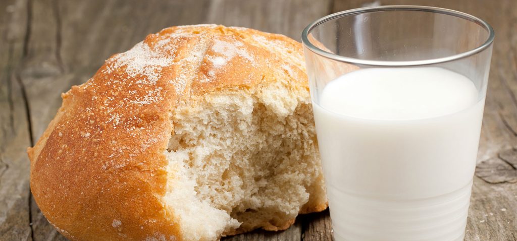 Milch mit Semmel Ein Glas Milch mit einem angebrochenen Brötchen in Nahaufnahme. - Natasha Breen/Shutterstock.com