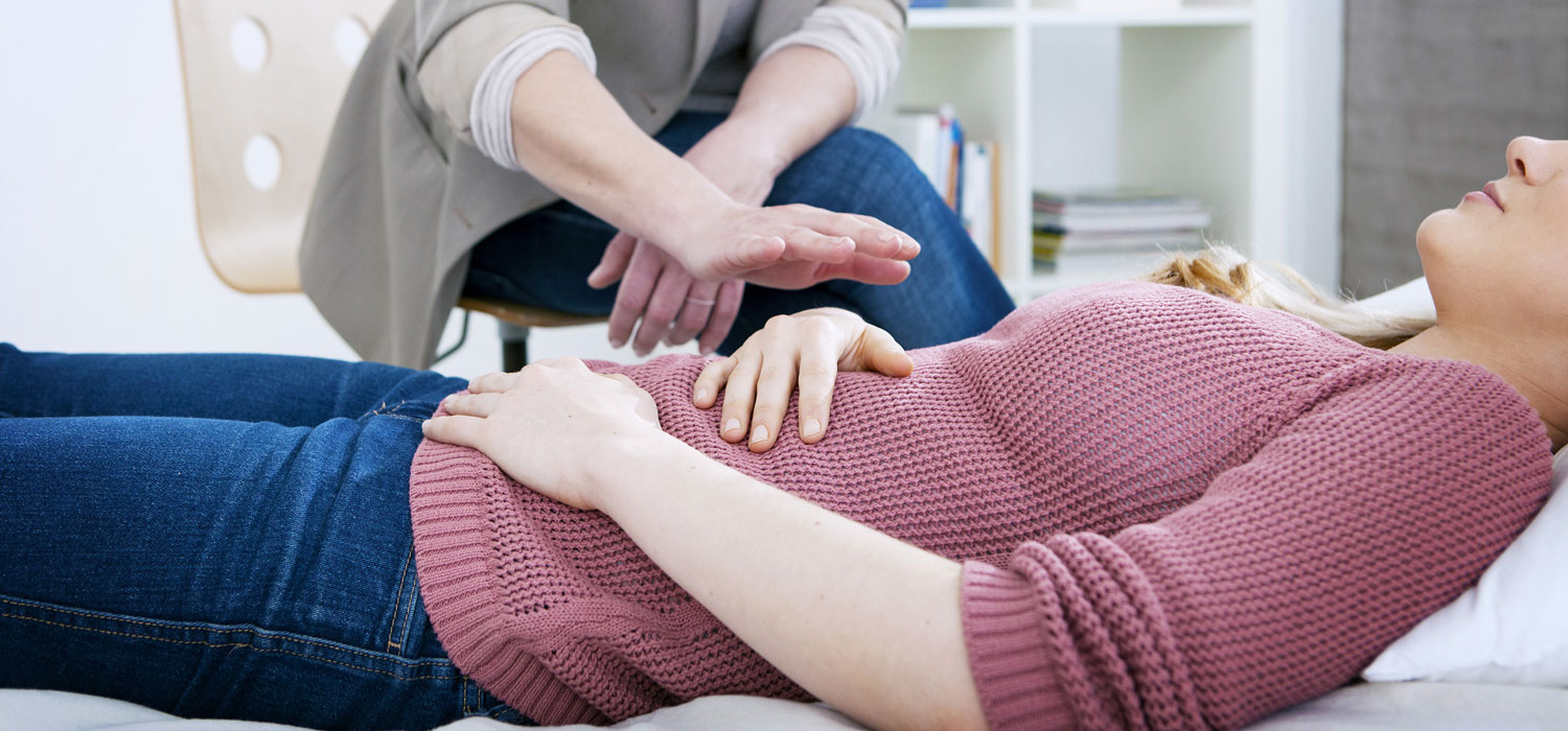 Patientin liegt am Rücken entspannt auf einem Behandlungsbett, die Therapeutin sitzt neben ihr. - Image Point Fr/Shutterstock.com