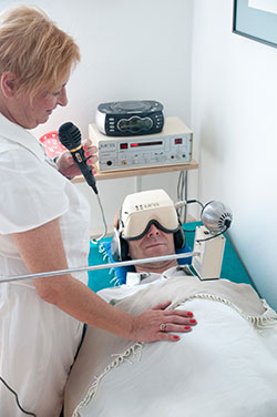 Patient am Rücken liegend mit Entspannungs- und Trance-Zustand fördernder Speziall-Brille und lärmdämmenden Kopfhörern. Die Hypnose-Ärztin steht neben ihm mit Mikro, sodass er nur ihre Stimme hören kann. - Jamina Paukner/Pumperlgsund