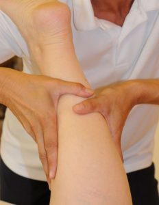 Lymphdrainage-Behandlung Nahaufnahme der Waden eines am Behandlungsbett liegenden Patienten; die Hände des Therapeuten streichen mit sanftem Druck in Richtung Herz. - Jamina Paukner Pumperlgsund