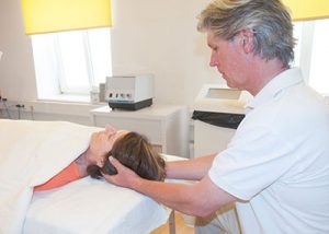 Masseur bei Behandlung Patientin in Behandlungsraum auf einem Massagebett am Rücken liegend; der Masseur steht zur Schädel-Nacken-Massage hinter hier. - Jamina Paukner/pumperlgsund