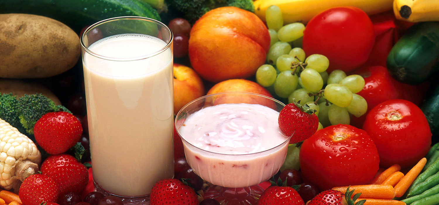 Stilleben-Foto mit zahlreichem frischem, vitaminreichem Obst und Gemüse sowie einem Glas Milch und einer Schale Erdbeer-Yoghurt.